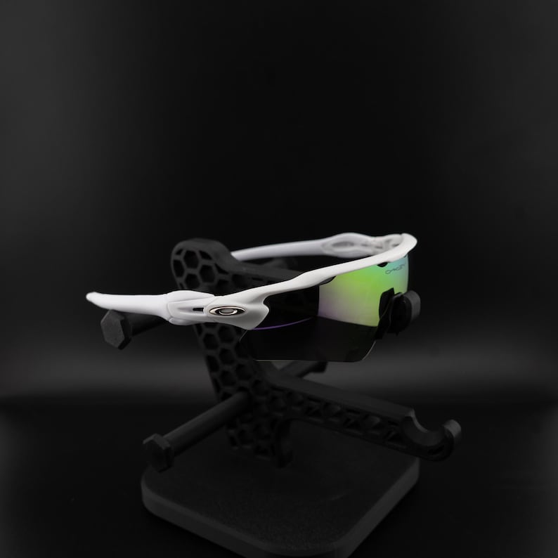 May include: White sports sunglasses with iridescent lenses are displayed on a black stand. The sunglasses have a sleek, modern design with a white frame and a prominent logo on the side. The lenses reflect a mix of green and purple hues.