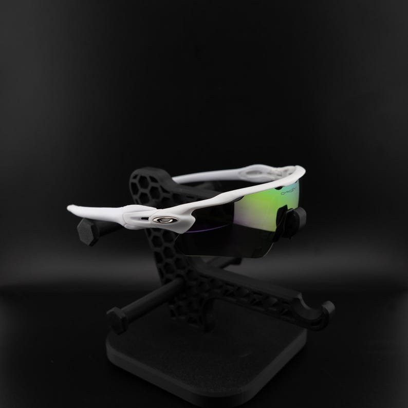 May include: White sports sunglasses with a reflective green and purple lens. The sunglasses are displayed on a black stand. The frame features the Oakley logo. The overall design is sleek and modern.