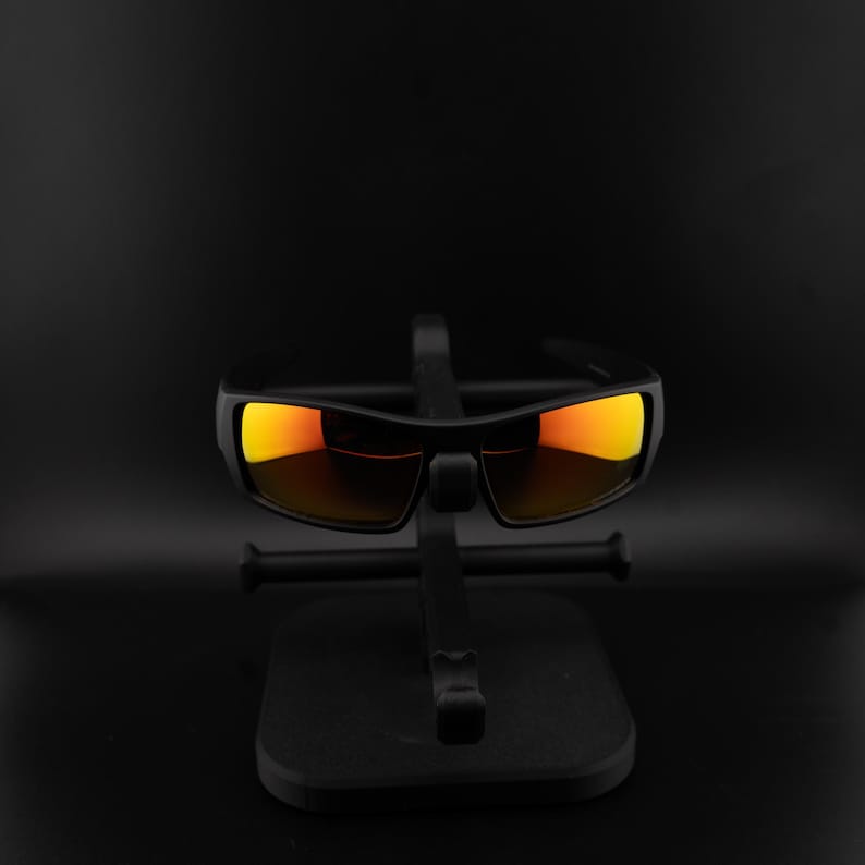 Pode incluir: &Oacute;culos de sol pretos com lentes laranja s&atilde;o exibidos em um suporte preto. Os &oacute;culos de sol retangulares t&ecirc;m uma arma&ccedil;&atilde;o grossa. As lentes refletem um tom laranja brilhante, contrastando com a arma&ccedil;&atilde;o e o suporte escuros. O suporte tamb&eacute;m &eacute; preto.