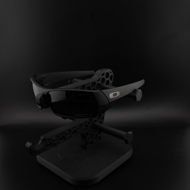 May include: A pair of black sunglasses with dark lenses rests on a black stand. The sunglasses have a rectangular frame and a modern design, with a small silver detail on the side. The stand has a geometric pattern.