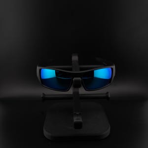 May include: Black sunglasses with blue mirrored lenses are displayed on a black stand. The sunglasses have a rectangular shape and a thick frame. The lenses reflect a bright blue color, contrasting with the dark frame and stand.