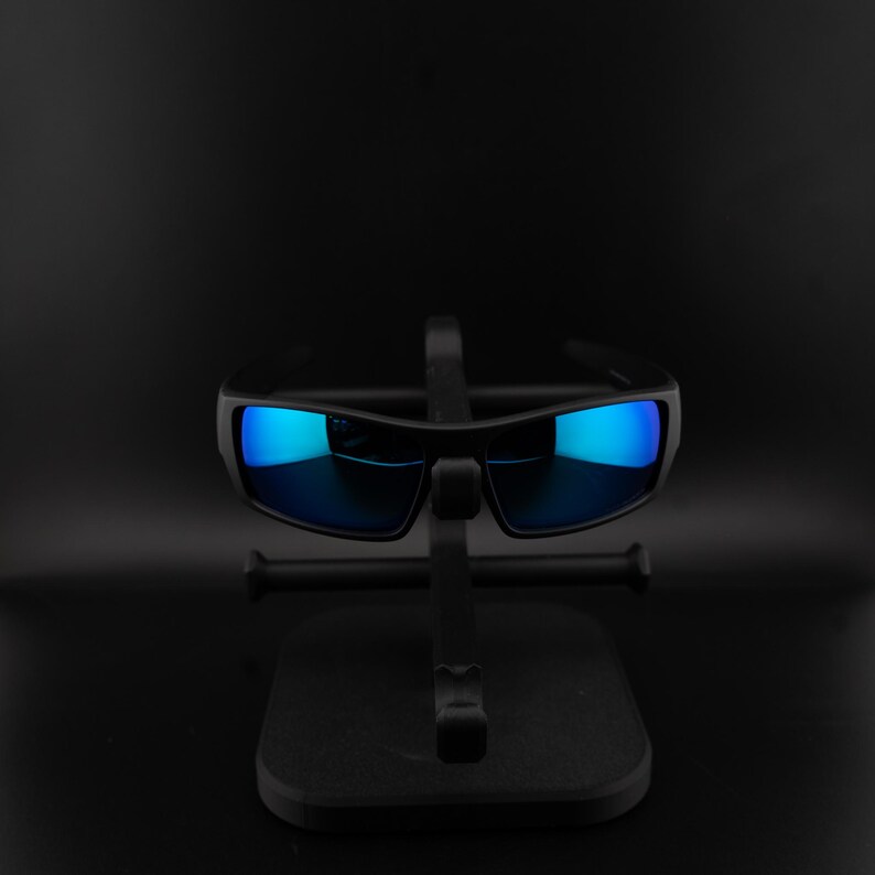 May include: Black sunglasses with blue mirrored lenses are displayed on a black stand. The sunglasses have a rectangular shape and a thick frame. The lenses reflect a bright blue color, contrasting with the dark frame and stand.