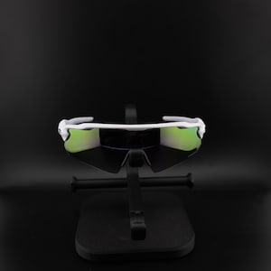May include: White sports sunglasses with green mirrored lenses. The sunglasses have a sleek, modern design and are displayed on a black stand against a black background. The lenses reflect a green hue.
