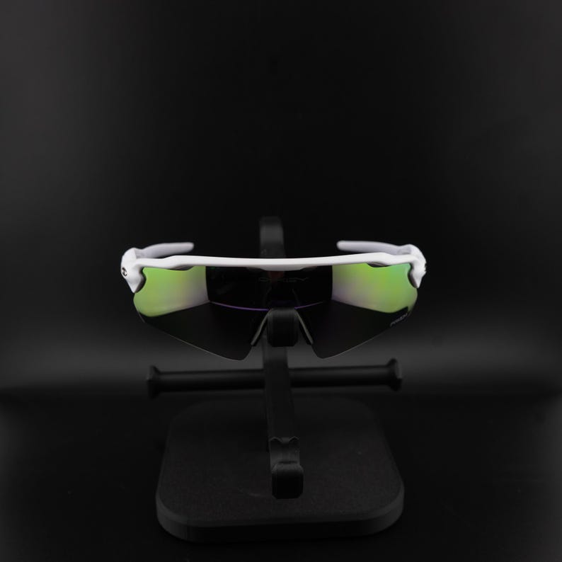 May include: White sports sunglasses with green mirrored lenses. The sunglasses have a sleek, modern design and are displayed on a black stand against a black background. The lenses reflect a green hue.