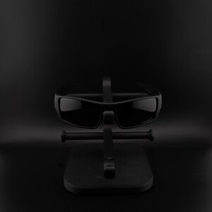 May include: Black sunglasses with a rectangular frame are displayed on a black stand. The sunglasses have dark lenses and a sleek, modern design. The stand is also black, providing a minimalist presentation.