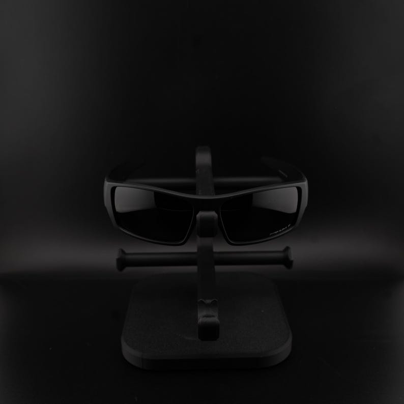 May include: Black sunglasses with a rectangular frame are displayed on a black stand. The sunglasses have dark lenses and a sleek, modern design. The stand is also black, providing a minimalist presentation.
