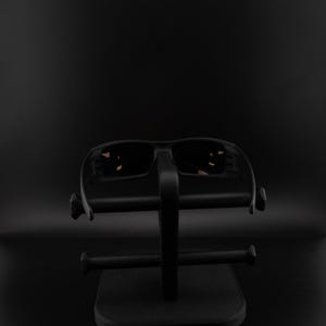 Pode incluir: &Oacute;culos de sol pretos com lentes escuras em um suporte de exibi&ccedil;&atilde;o preto. Os &oacute;culos de sol t&ecirc;m um design esportivo, e o suporte tem uma base retangular com um suporte vertical e bra&ccedil;os horizontais.