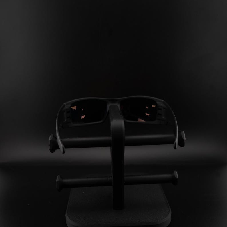 Pode incluir: &Oacute;culos de sol pretos com lentes escuras em um suporte de exibi&ccedil;&atilde;o preto. Os &oacute;culos de sol t&ecirc;m um design esportivo, e o suporte tem uma base retangular com um suporte vertical e bra&ccedil;os horizontais.