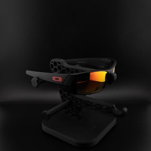 Pode incluir: &Oacute;culos de sol pretos com lentes espelhadas laranja s&atilde;o exibidos em um suporte preto. Os &oacute;culos de sol de arma&ccedil;&atilde;o retangular apresentam um logotipo vermelho. O suporte tem um design geom&eacute;trico e uma base quadrada.