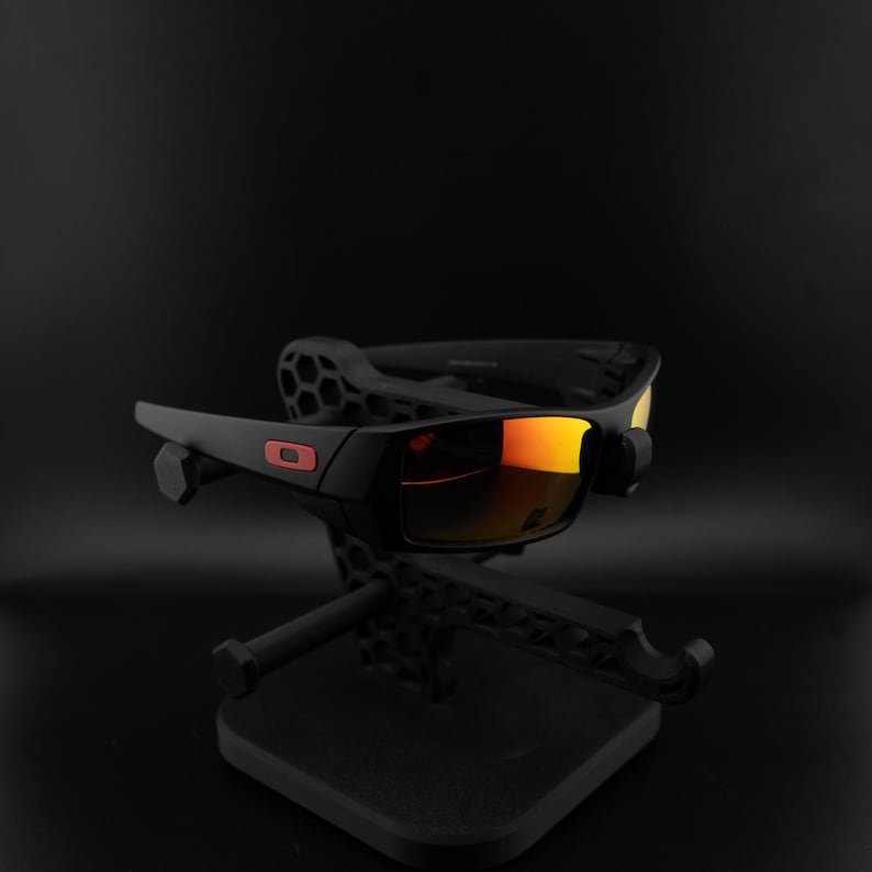 Pode incluir: &Oacute;culos de sol pretos com lentes espelhadas laranja em um suporte preto. Os &oacute;culos t&ecirc;m formato retangular com um logotipo vermelho. O suporte apresenta um design geom&eacute;trico.