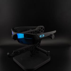 May include: Black sunglasses with blue mirrored lenses are displayed on a black stand. The sunglasses have a sleek, modern design with a small blue accent on the frame. The stand has a geometric design.
