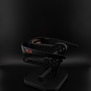Pode incluir: &Oacute;culos de sol pretos com um detalhe vermelho na arma&ccedil;&atilde;o s&atilde;o exibidos em um suporte preto. O suporte tem um design geom&eacute;trico e uma base quadrada. O fundo &eacute; preto liso.