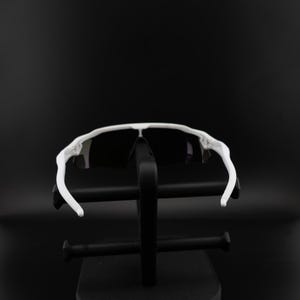 May include: White sports sunglasses with black lenses. The sunglasses have a sleek, modern design with a white frame and curved temples. The sunglasses are displayed on a black stand against a black background.
