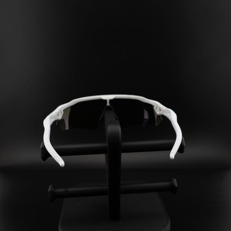 May include: White sports sunglasses with black lenses. The sunglasses have a sleek, modern design with a white frame and curved temples. The sunglasses are displayed on a black stand against a black background.