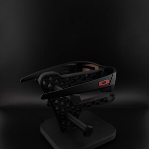 Pode incluir: &Oacute;culos de sol pretos com um detalhe vermelho na haste, expostos em um suporte preto. Os &oacute;culos t&ecirc;m um design moderno e elegante. O suporte apresenta um padr&atilde;o geom&eacute;trico e uma base quadrada.
