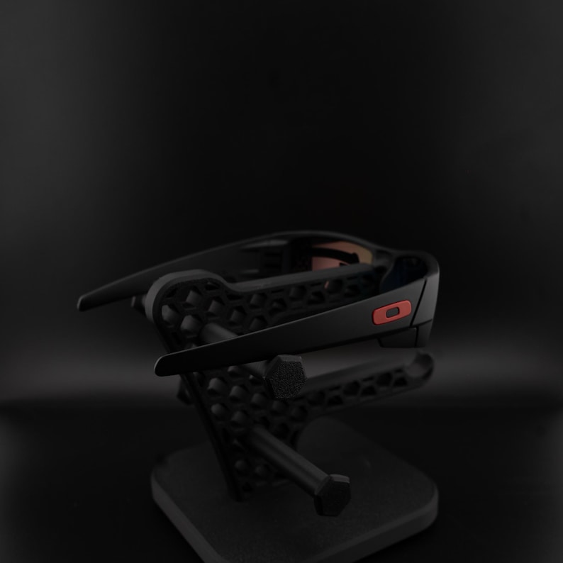 Pode incluir: &Oacute;culos de sol pretos com um detalhe vermelho na haste, expostos em um suporte preto. Os &oacute;culos t&ecirc;m um design moderno e elegante. O suporte apresenta um padr&atilde;o geom&eacute;trico e uma base quadrada.