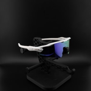 May include: White-framed sunglasses with blue and green lenses rest on a black display stand. The sunglasses have a sleek, modern design. The stand is also black and has a geometric pattern.