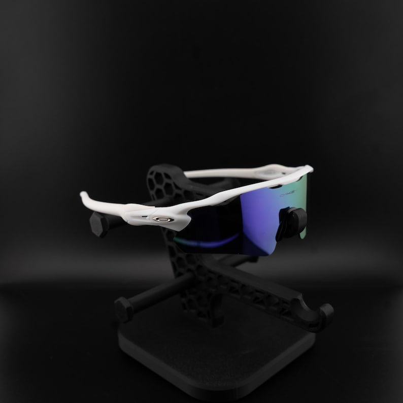 May include: White-framed sunglasses with blue and green lenses rest on a black display stand. The sunglasses have a sleek, modern design. The stand is also black and has a geometric pattern.