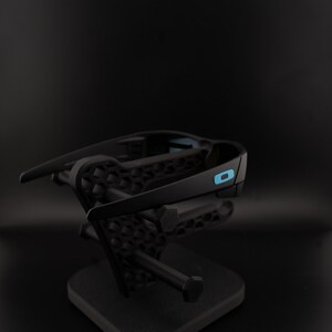 May include: Black sunglasses with a blue accent on the arm are displayed on a black stand. The sunglasses have a sleek, modern design. The stand has a geometric pattern and a sturdy base, providing a stylish presentation.