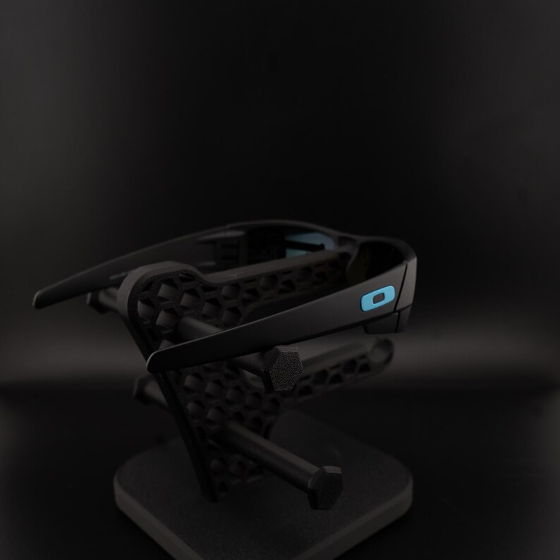 May include: Black sunglasses with a blue accent on the arm are displayed on a black stand. The sunglasses have a sleek, modern design. The stand has a geometric pattern and a sturdy base, providing a stylish presentation.