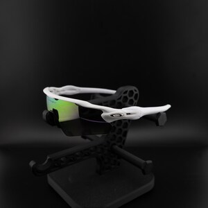 May include: White sports sunglasses with a curved frame and iridescent lenses. The sunglasses are displayed on a black stand with a geometric design. The Oakley logo is visible on the temple.