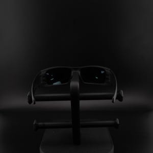 May include: Black sunglasses with dark lenses rest on a black display stand. The sunglasses have a rectangular shape and a thick frame. The stand has a minimalist design with horizontal bars.