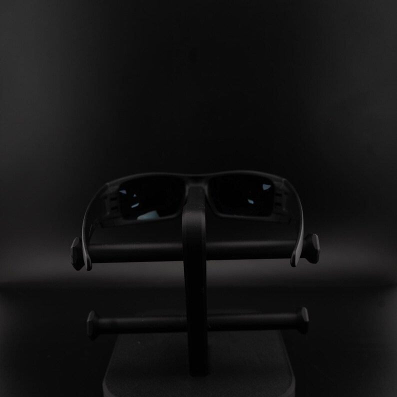 May include: Black sunglasses with dark lenses rest on a black display stand. The sunglasses have a rectangular shape and a thick frame. The stand has a minimalist design with horizontal bars.