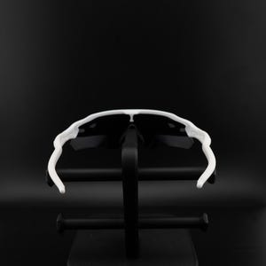 May include: White-framed sunglasses with black lenses are displayed on a black stand against a black background. The sunglasses have a sporty design with a curved frame and arms. The lenses are dark, providing contrast to the white frame.