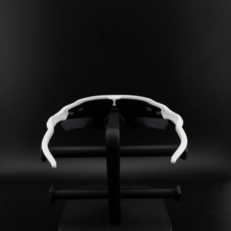 May include: White-framed sunglasses with black lenses are displayed on a black stand against a black background. The sunglasses have a sporty design with a curved frame and arms. The lenses are dark, providing contrast to the white frame.