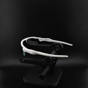 May include: White sports sunglasses with a black frame and a black stand. The sunglasses have a sleek, modern design with a curved frame and dark lenses. The stand is a geometric design.