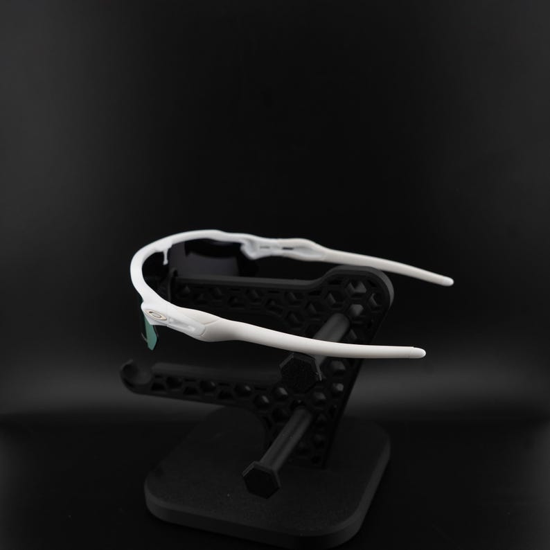 May include: White sports sunglasses with a black frame and a black stand. The sunglasses have a sleek, modern design with a curved frame and dark lenses. The stand is a geometric design.
