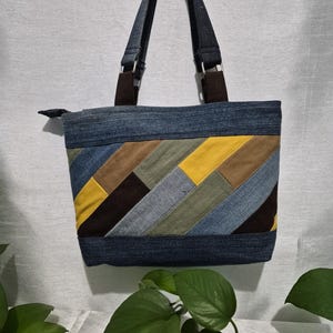 May include: A denim tote bag with a patchwork design featuring yellow, brown, and blue panels. The bag has dark blue denim handles and a zipper closure. The bag is hanging against a white wall.
