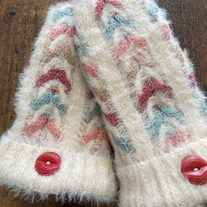 Puede incluir: Un par de mitones de punto color crema con un patrón texturizado en rosa, azul y marrón claro. Cada mitón tiene un botón rojo y una textura esponjosa y difusa.