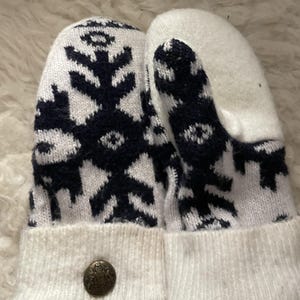 Puede incluir: Un par de mitones blancos con un diseño de copo de nieve azul marino. Los puños son blancos y tienen un botón decorativo. Los mitones están hechos de un material de punto suave, ideal para el frío.