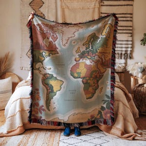 May include: A rectangular throw blanket featuring a vintage world map design. The map is in shades of green, brown, and yellow, with a light blue background. The blanket has a fringed edge with red, black, and brown accents. The text "ELLED ARE 'CIROPTIN'" is visible.