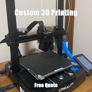 May include: A black 3D printer with the text "Custom 3D Printing" and "Free Quote". The printer has a black base, a black print bed, and a black and silver print head. A small screen is visible on the right side.