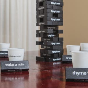 Puede incluir: Un juego "Tipsy Tower" negro con vasos de chupito blancos y pequeños bloques negros con frases como "give one take one" y "rhyme time". La torre está apilada sobre una mesa de madera.