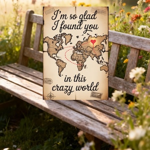 Puede incluir: Una impresión de mapa del mundo de estilo vintage con el texto "I'm so glad I found you in this crazy world". El mapa presenta un corazón rojo y líneas punteadas que conectan diferentes ubicaciones. La impresión se muestra en un banco de madera.
