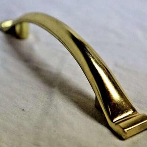May include: A gold-toned metal cabinet pull with a curved design. The pull has a smooth, polished finish.