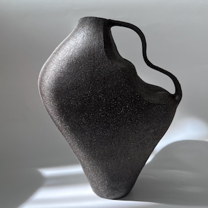 Puede incluir: Un jarrón de cerámica de color carbón oscuro con una forma asimétrica única. El jarrón tiene una superficie texturizada y un asa curva. El diseño es moderno y artístico, adecuado para la decoración del hogar.