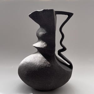 Può includere: Un vaso in ceramica nera dal design astratto. Il vaso ha una base bulbosa, un collo scolpito con un elemento sporgente e un manico a forma di zigzag ondulato. La superficie ha una finitura opaca e strutturata.