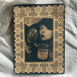 May include: A rectangular print featuring a sepia-toned photograph of two people, one wearing glasses and a hat, framed by a decorative tan border with a repeating floral lace pattern against a dark background, creating a vintage aesthetic.