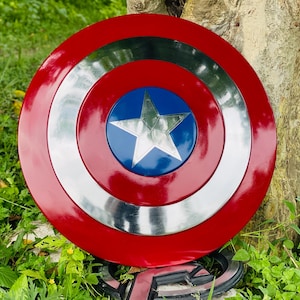 May include: A red, white, and blue shield with a silver star in the center. The shield has a circular design with concentric rings of red, silver, and blue. The shield is resting on green grass.