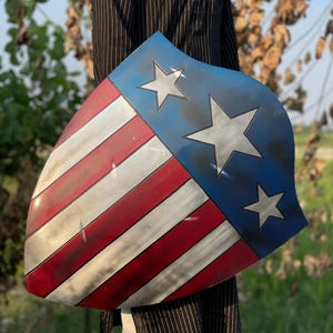 Op de afbeelding: Een schild met een patriottisch ontwerp, met rode en witte strepen en een blauw gedeelte met drie witte sterren. Het schild heeft een verweerde, verouderde uitstraling, met zichtbare slijtage.