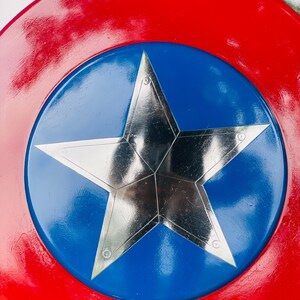 May include: A red, white, and blue shield with a large silver star in the center. The shield has a red outer ring, a blue inner circle, and a five-pointed star in the middle. The shield is a replica of a superhero's shield.