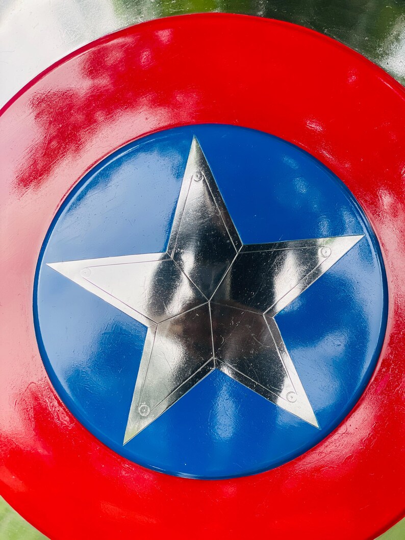 May include: A red, white, and blue shield with a large silver star in the center. The shield has a red outer ring, a blue inner circle, and a five-pointed star in the middle. The shield is a replica of a superhero's shield.