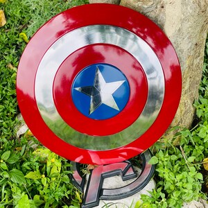 May include: A Captain America shield replica. The shield is round with a red outer ring, a silver middle ring, and a blue center with a white star. The Avengers logo is visible in the background.