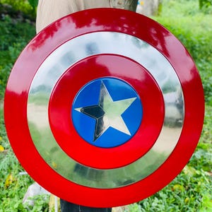 May include: A round, red and silver shield with a blue center featuring a silver star. The shield has a glossy finish and is held in front of a person. The shield is a replica of a superhero's shield.