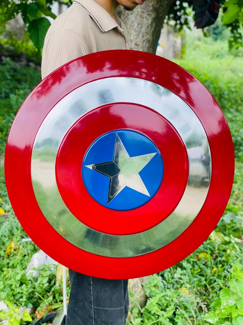 May include: A round, red and silver shield with a blue center featuring a silver star. The shield has a glossy finish and is held in front of a person. The shield is a replica of a superhero's shield.