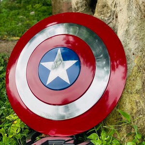 May include: A red, white, and blue Captain America shield. The shield has a circular design with a silver outer ring, a red inner ring, and a blue center with a white star. The shield is resting on a stone.